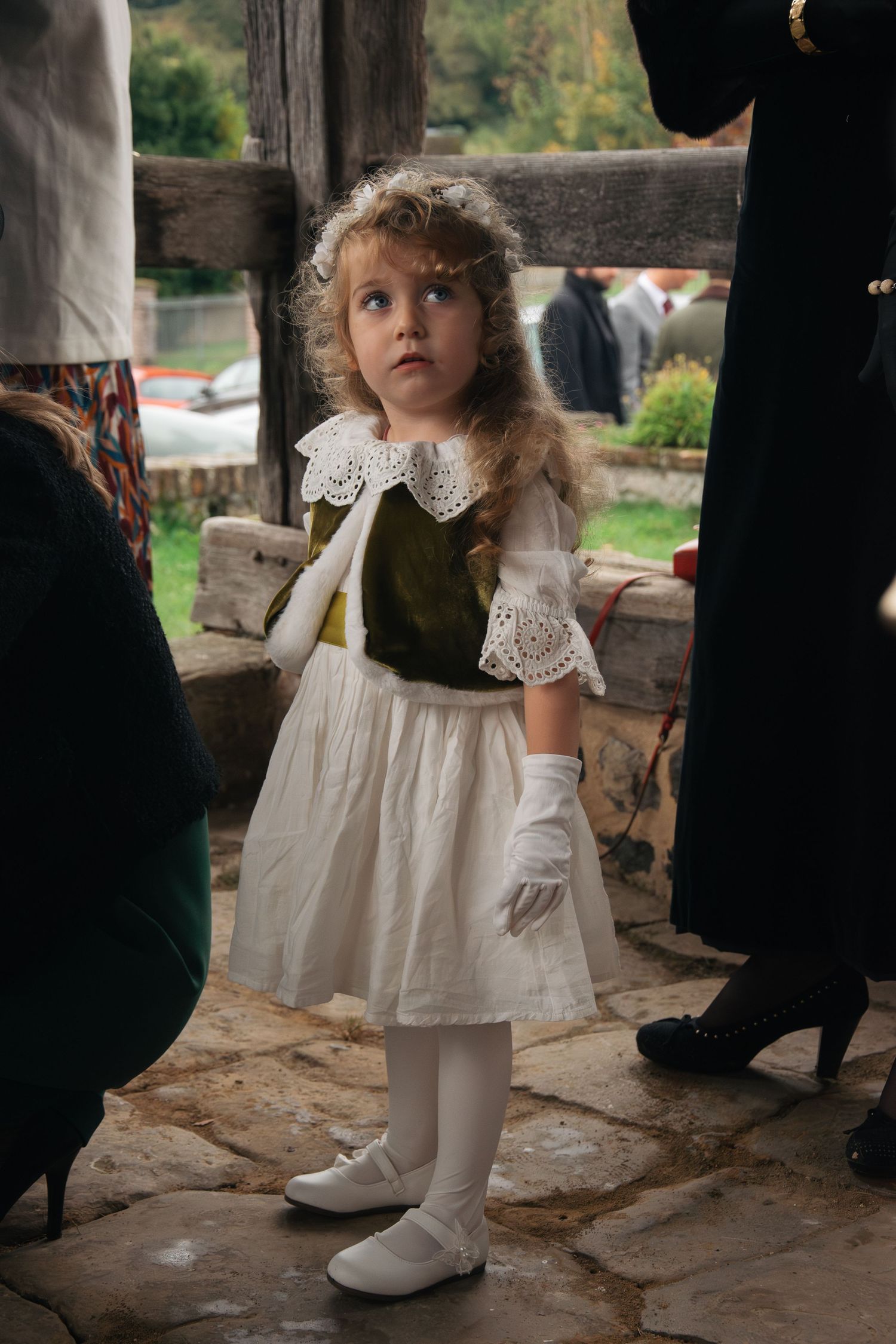 Young flower girl in vintage dress and lace collar stands on stone patio during wedding.