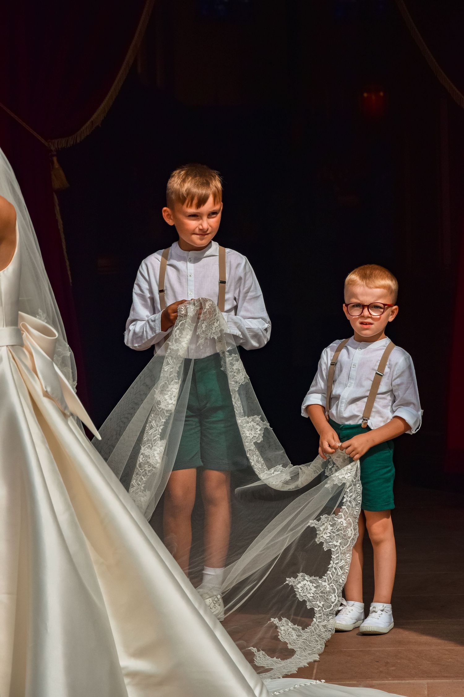 Two young pageboys in suspenders carrying the train of a wedding dress.