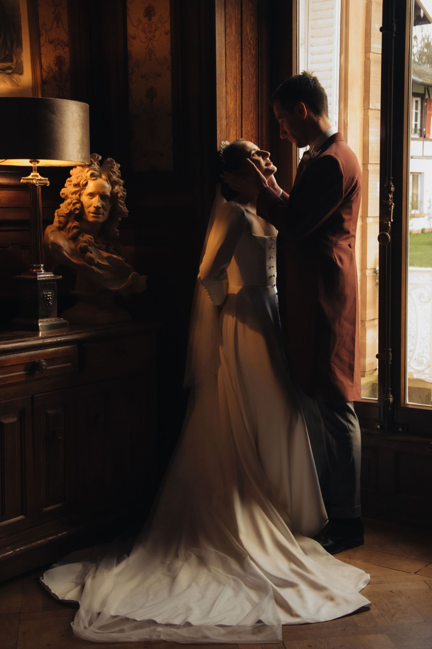 A couple shares an intimate moment in warm sunlight while standing by a window with a flowing white wedding dress.