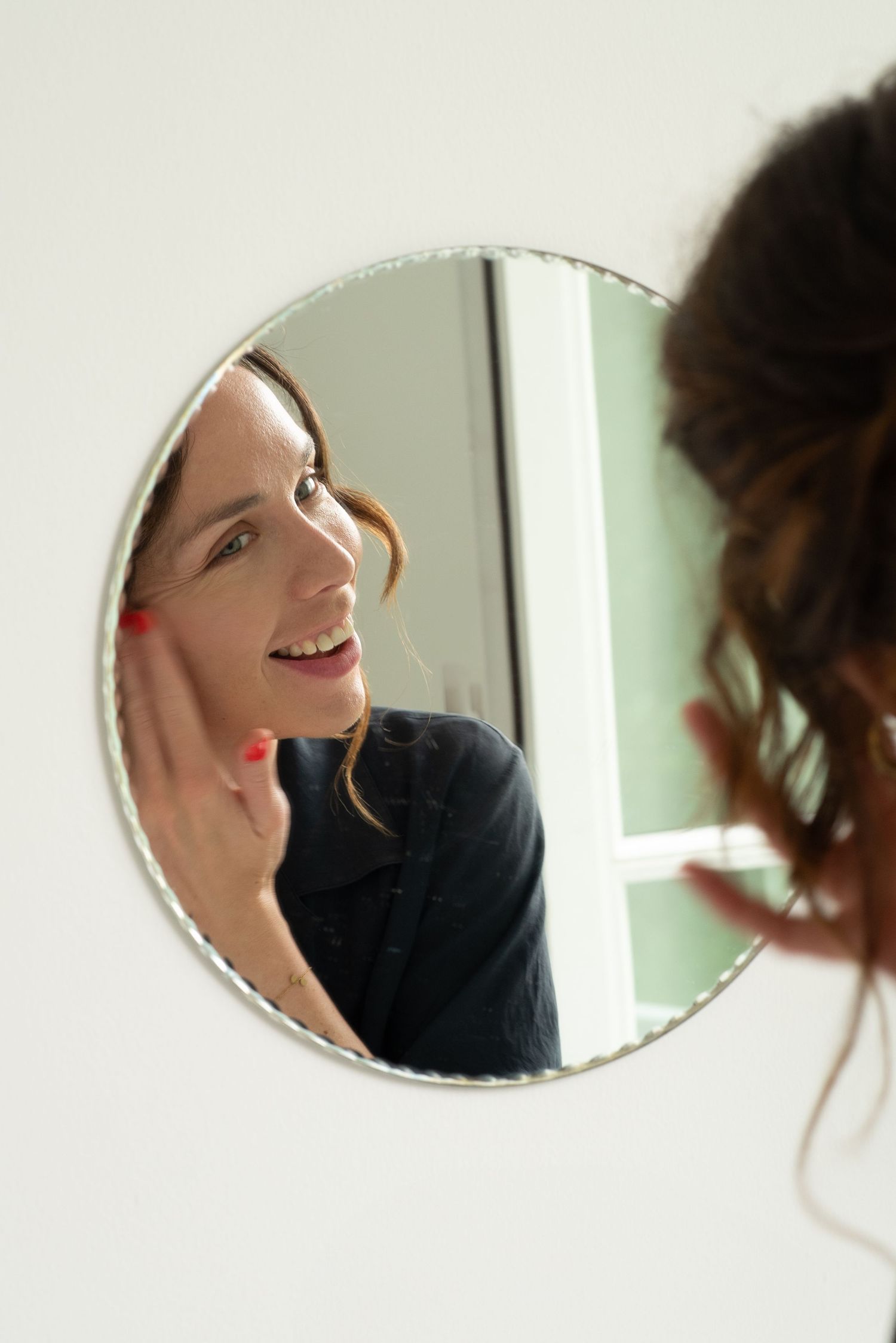A reflection in a circular mirror shows someone smiling while touching their face against a light colored wall.