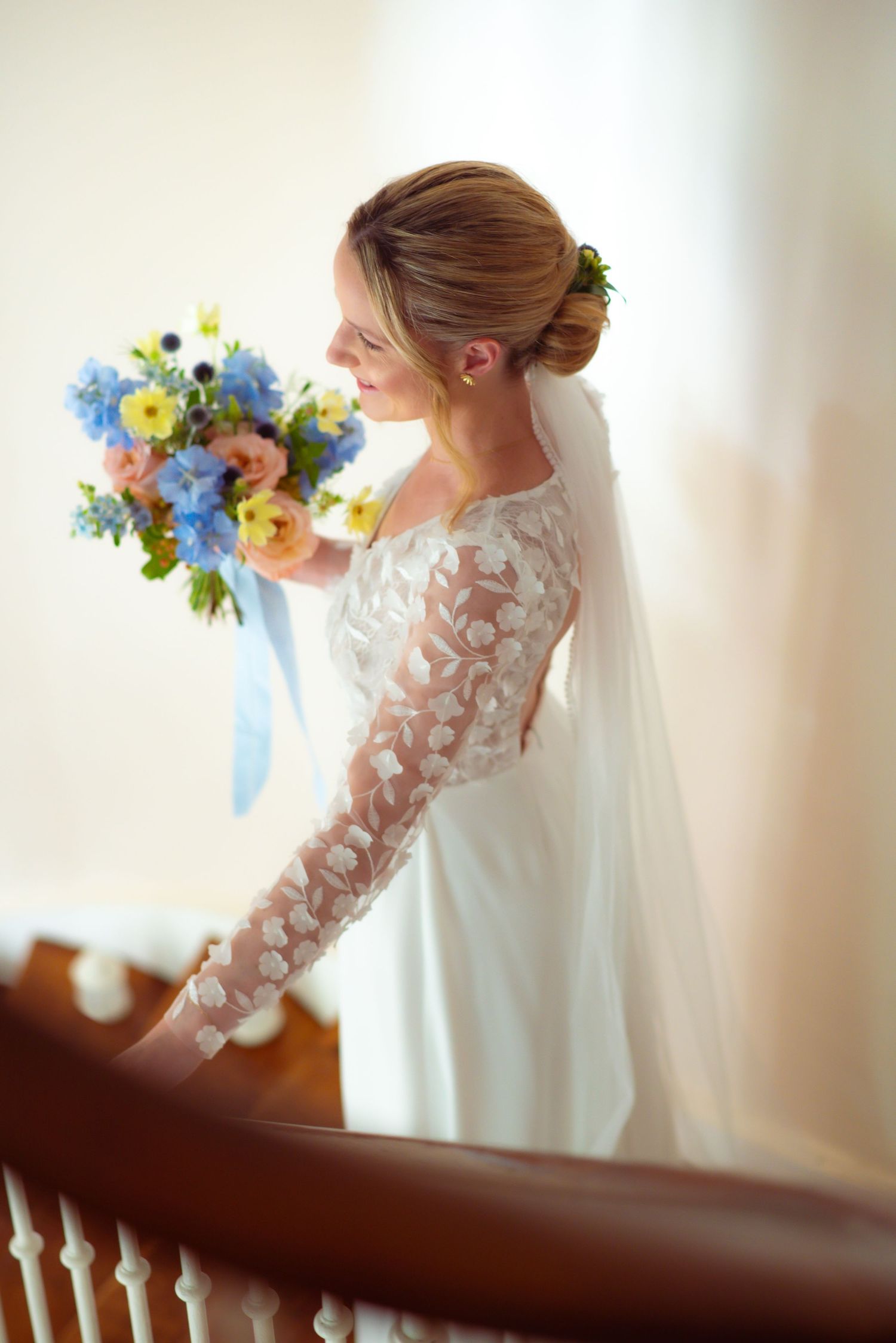 A bride in a lace sleeve wedding gown holds a vibrant blue and yellow bouquet while standing by wooden banister.
