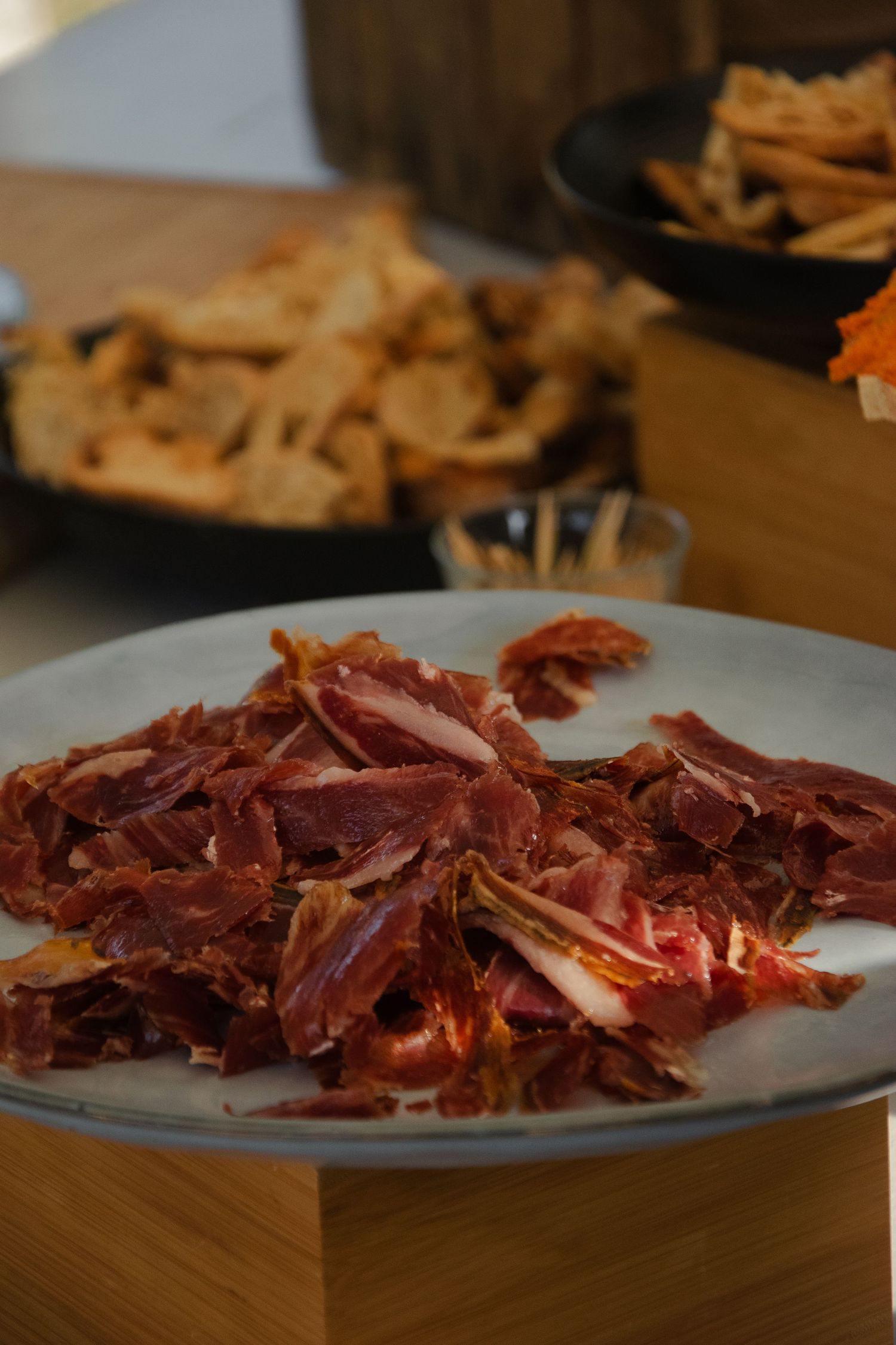 Thin slices of cured Spanish jamon iberico ham arranged on a white plate against a wooden table surface.