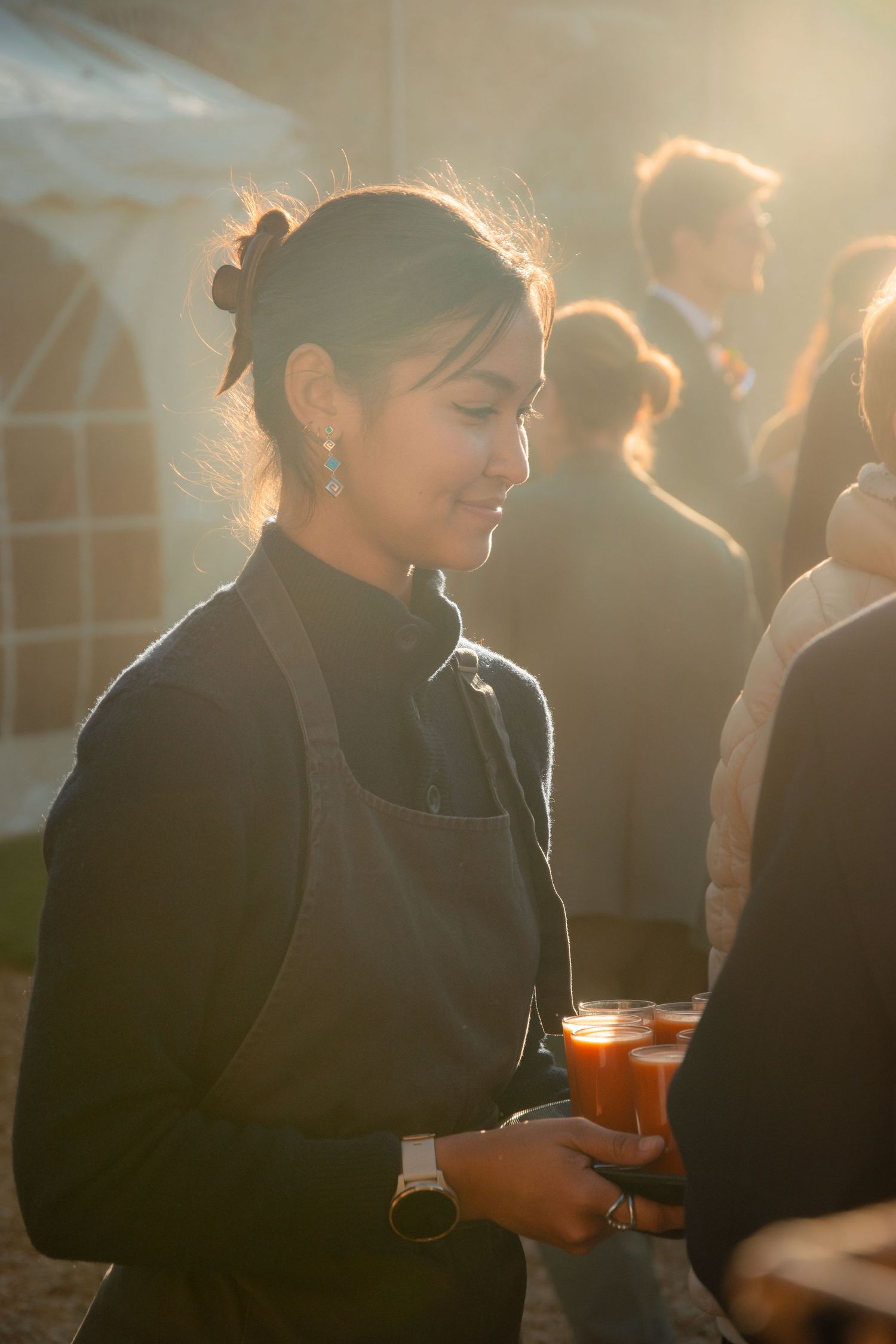 Server in black attire holding orange cocktail during sunset reception.