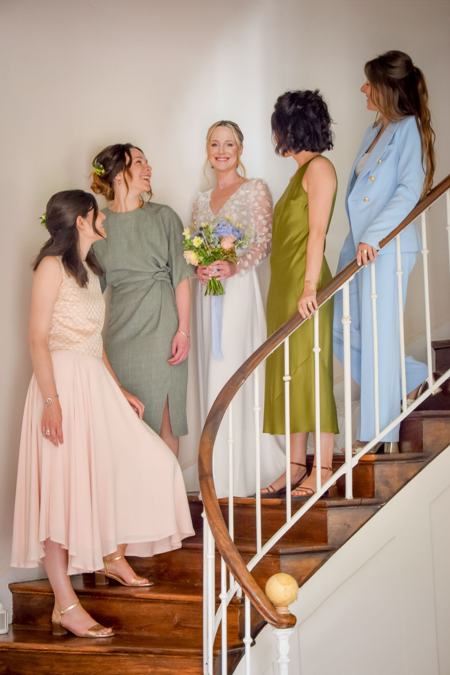 A bridal party in pastel and jewel-toned dresses gather on an elegant wooden staircase with white railings.