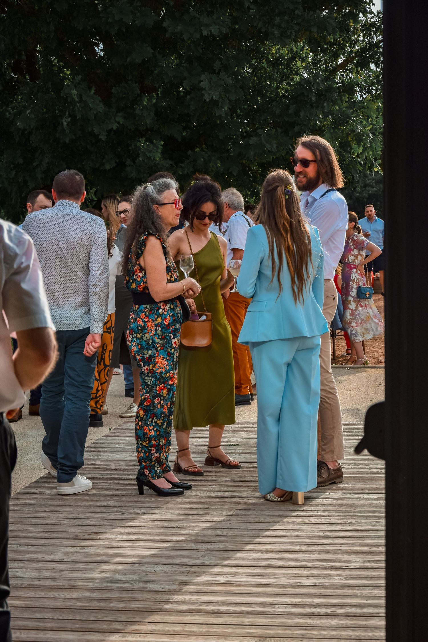 A group of people gathered on a wooden deck wearing colorful summer outfits during an outdoor social event.