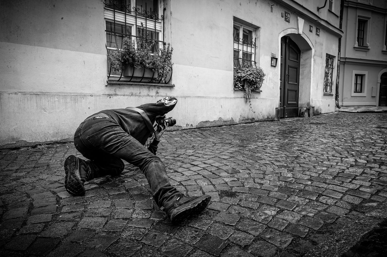 Black and white photo of a photographer lying down on a cobblestone street while taking a shot in an urban setting.