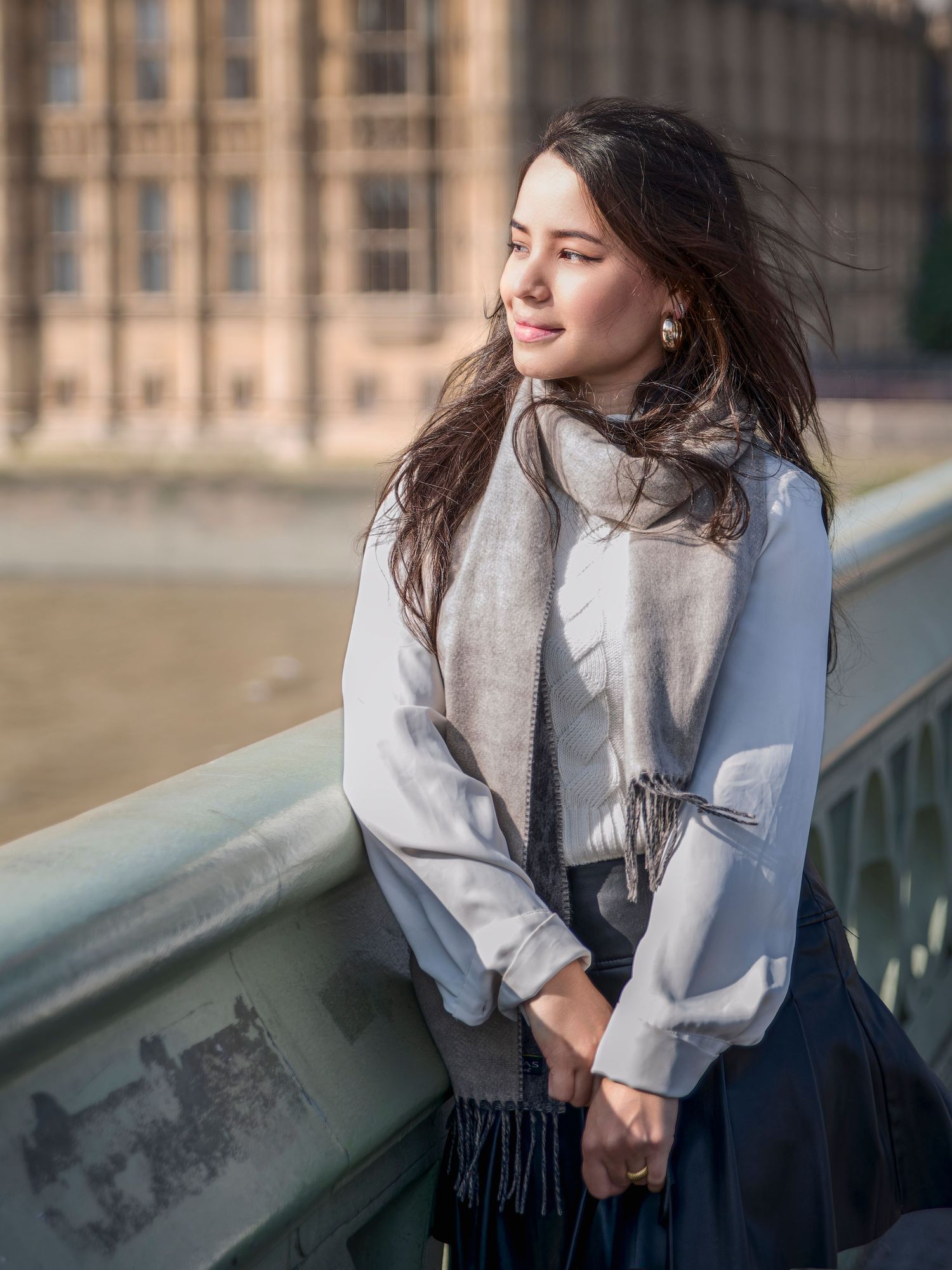 A person in a white shirt and dark skirt leans against a railing while looking away thoughtfully in an urban setting.