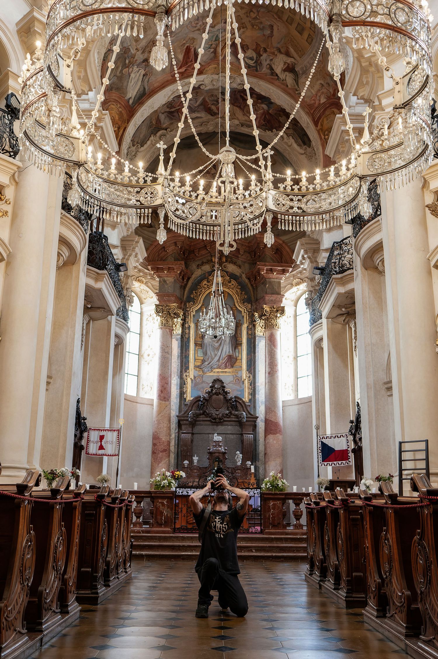 Stunning baroque church interior with ornate crystal chandelier, carved wooden pews, and person in black at center aisle.