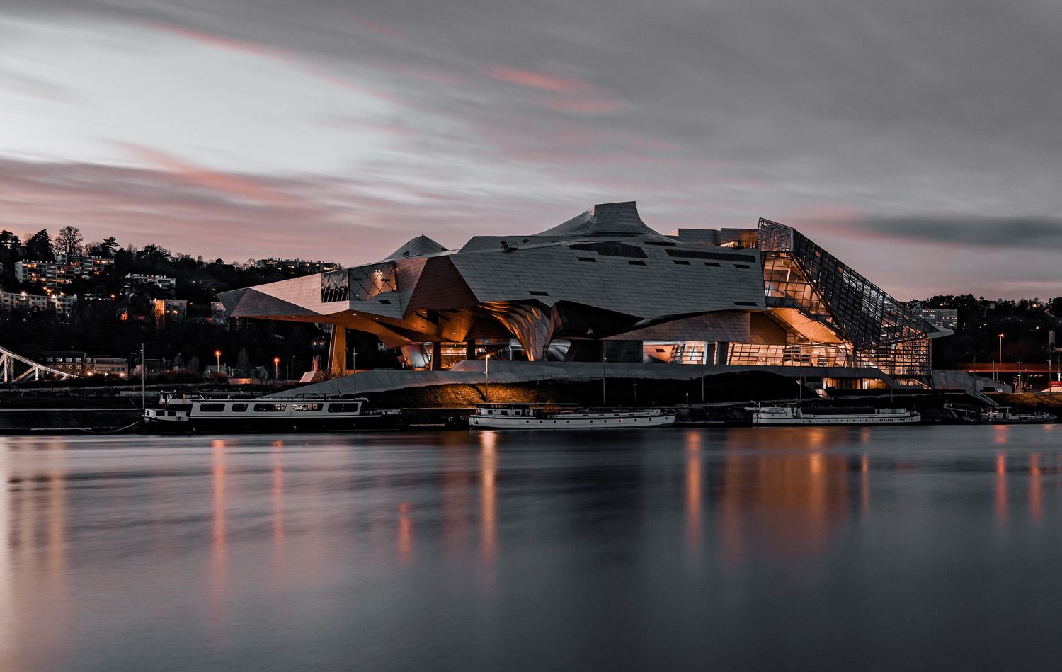 Modern architectural building illuminated at dusk reflecting on calm water under dramatic cloudy sky.