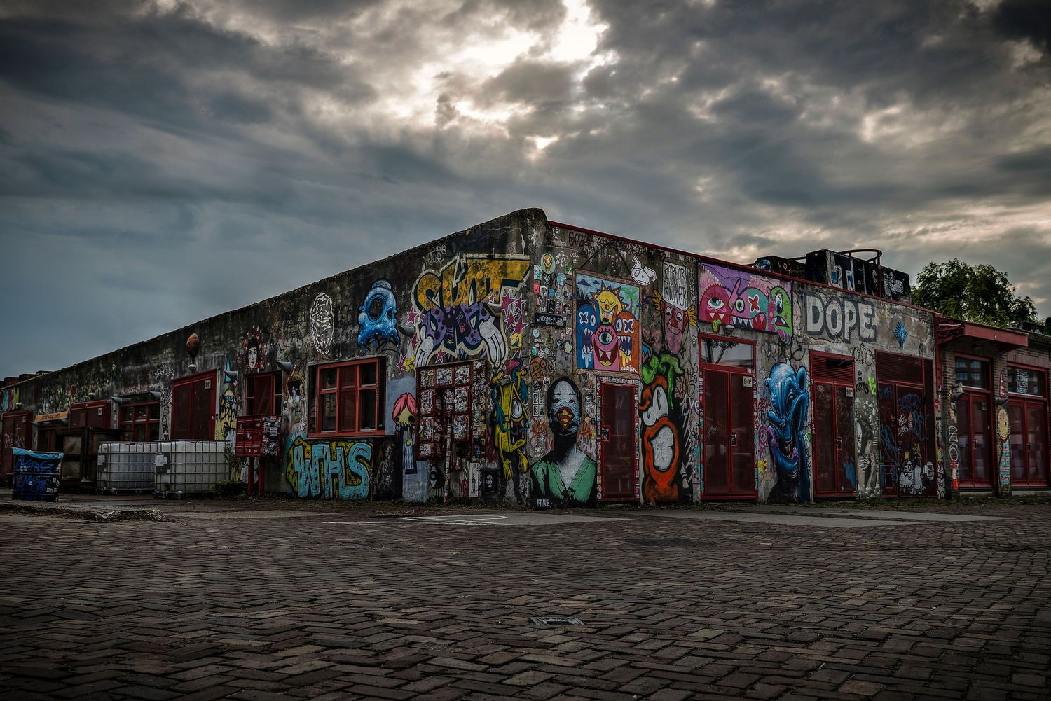 Old abandoned warehouse building covered in colorful graffiti art under dramatic cloudy sky at dusk.