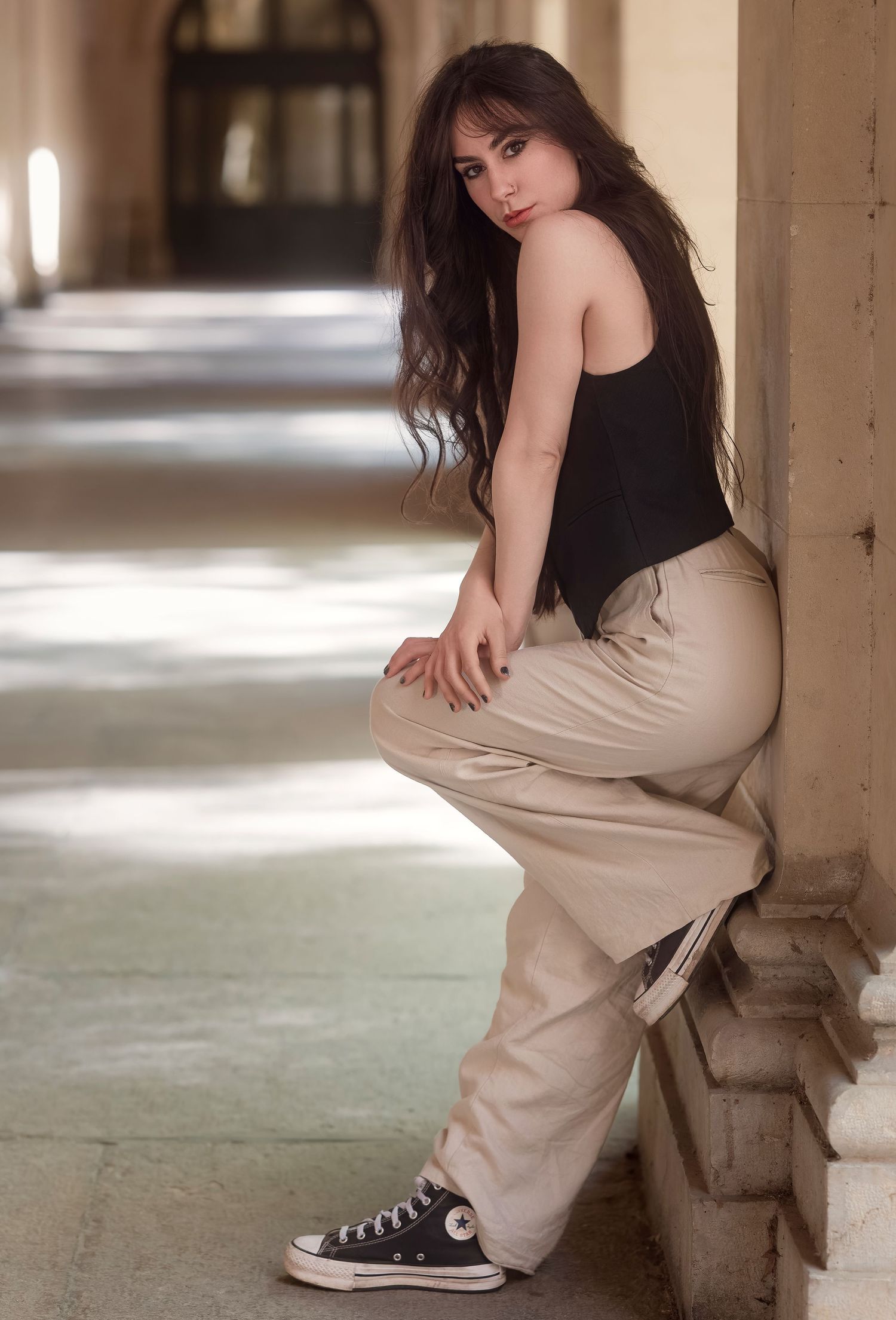 Young woman in black top, beige cargo pants, and Converse sneakers posing gracefully against a stone column.