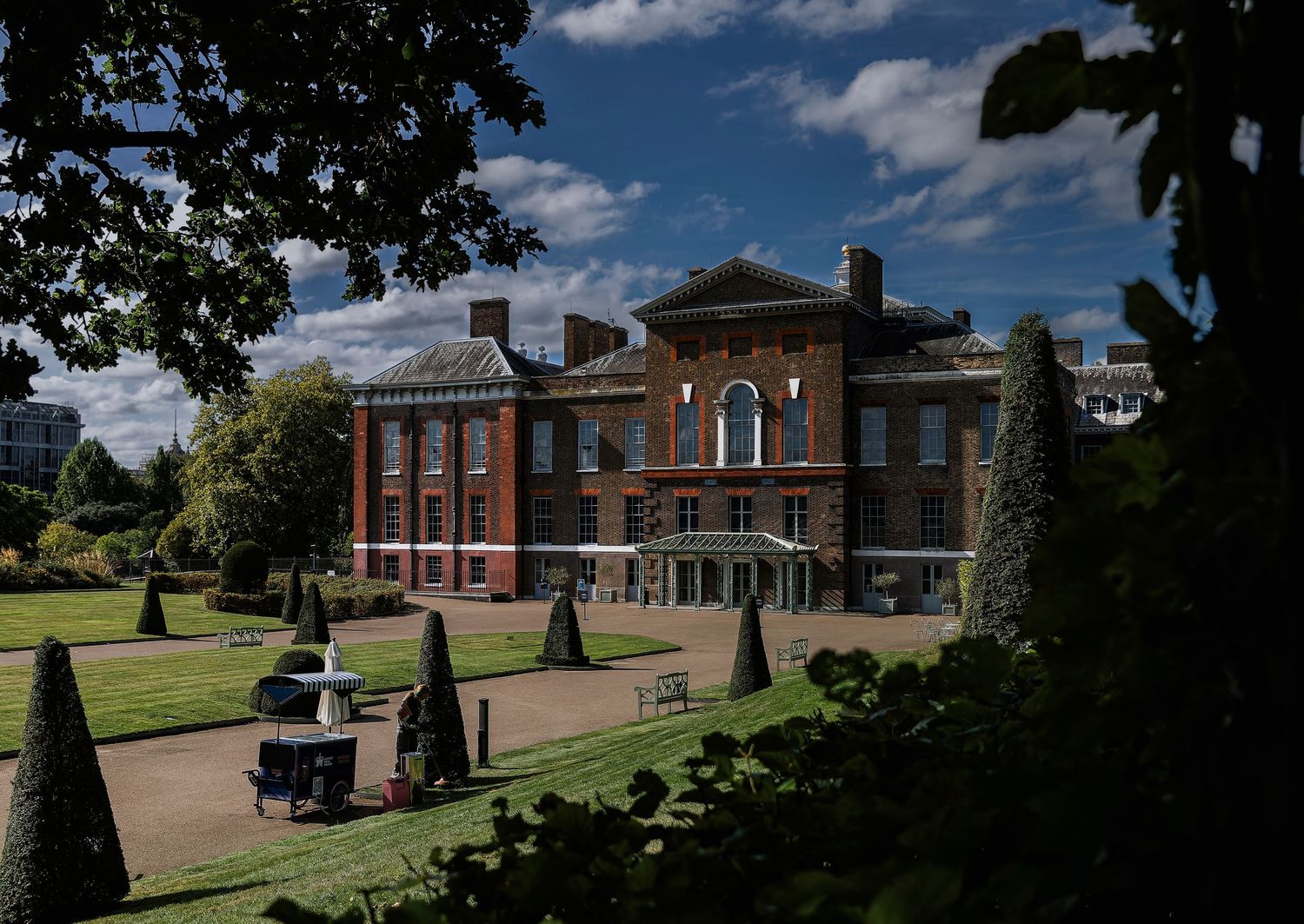 A stately red brick mansion with manicured gardens and topiary trees sits under a bright blue summer sky.