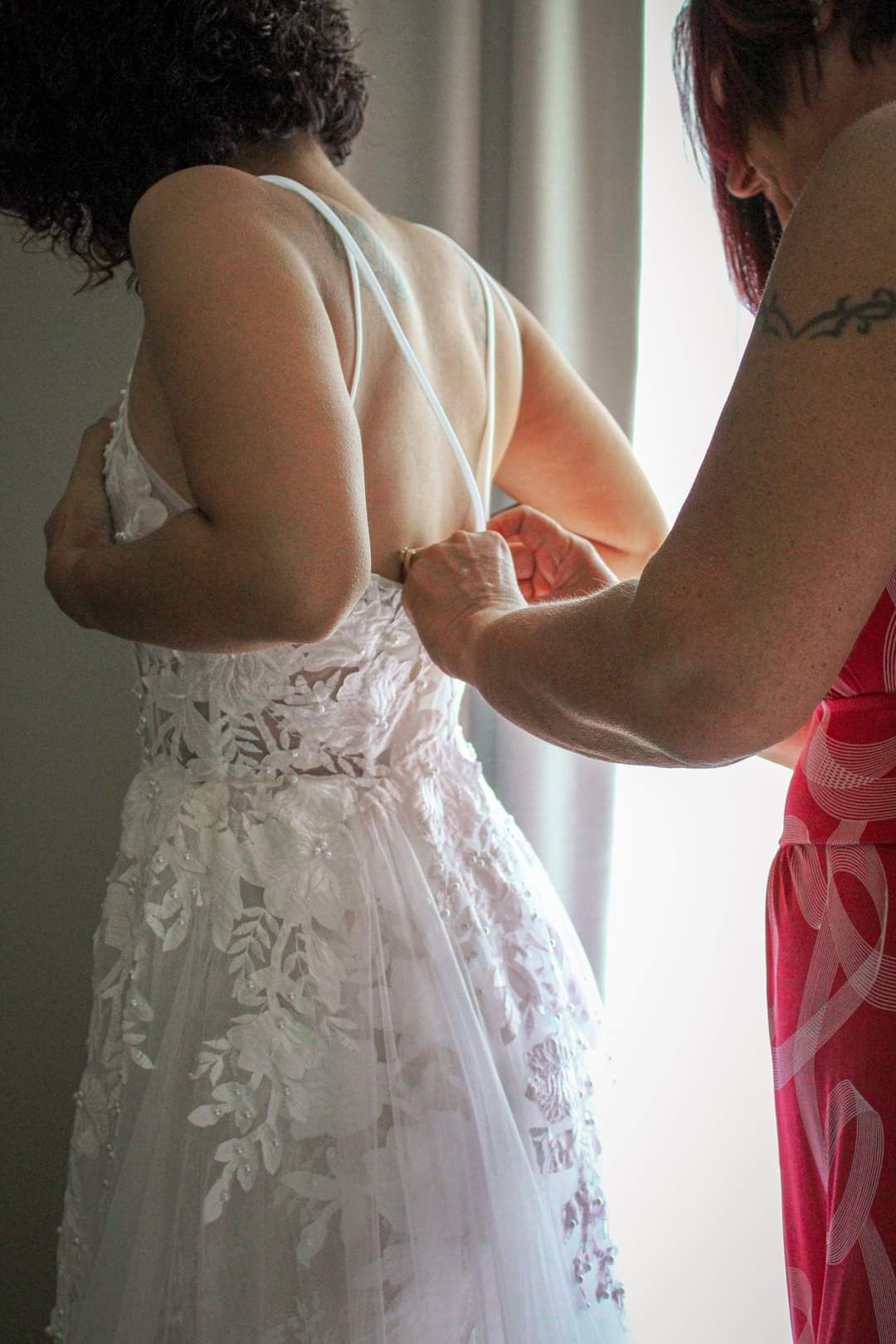 A bridesmaid in red helps button the back of a bride's white lace wedding dress near a bright window.