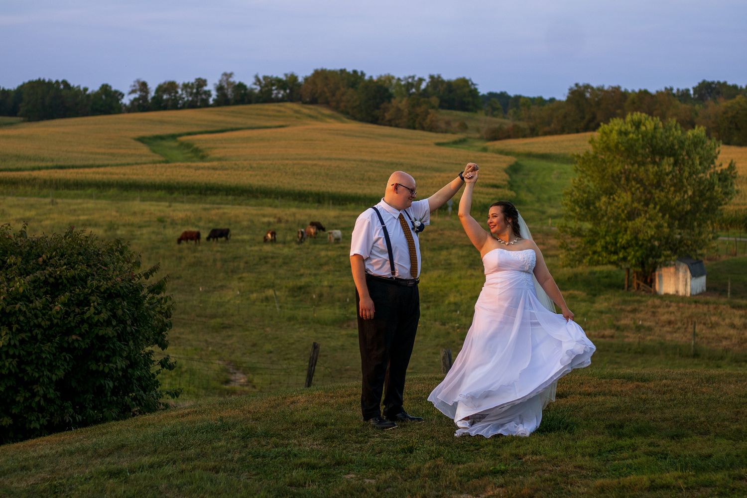 A couple dances together at sunset on a hillside farm with rolling pastures and hay bales in the background.