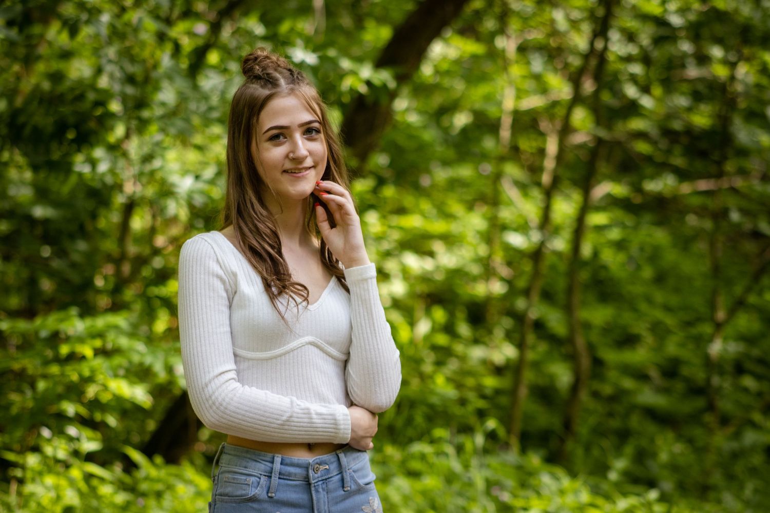 Young woman in a white sweater and jeans poses thoughtfully in front of lush green forest background.