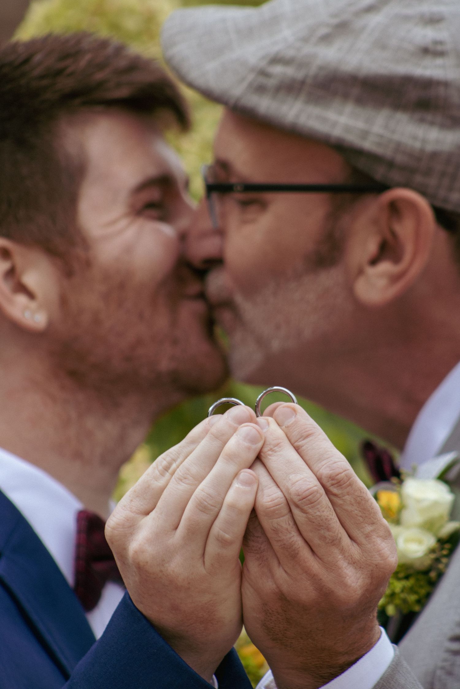 Close up of hands holding wedding rings as two people share a kiss, with a yellow flower boutonniere visible.