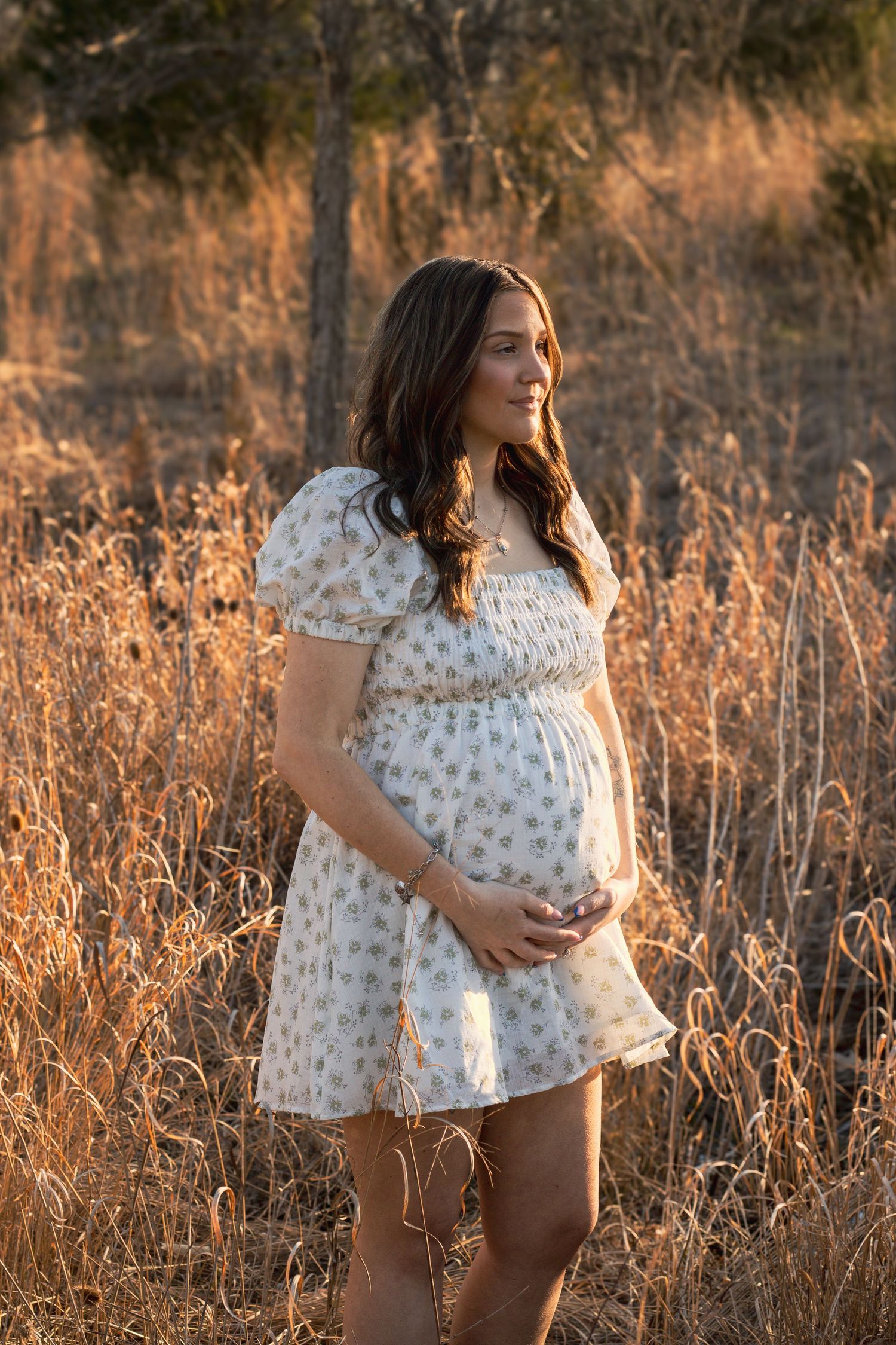 Pregnant woman in white floral dress holding baby bump in golden field at sunset.