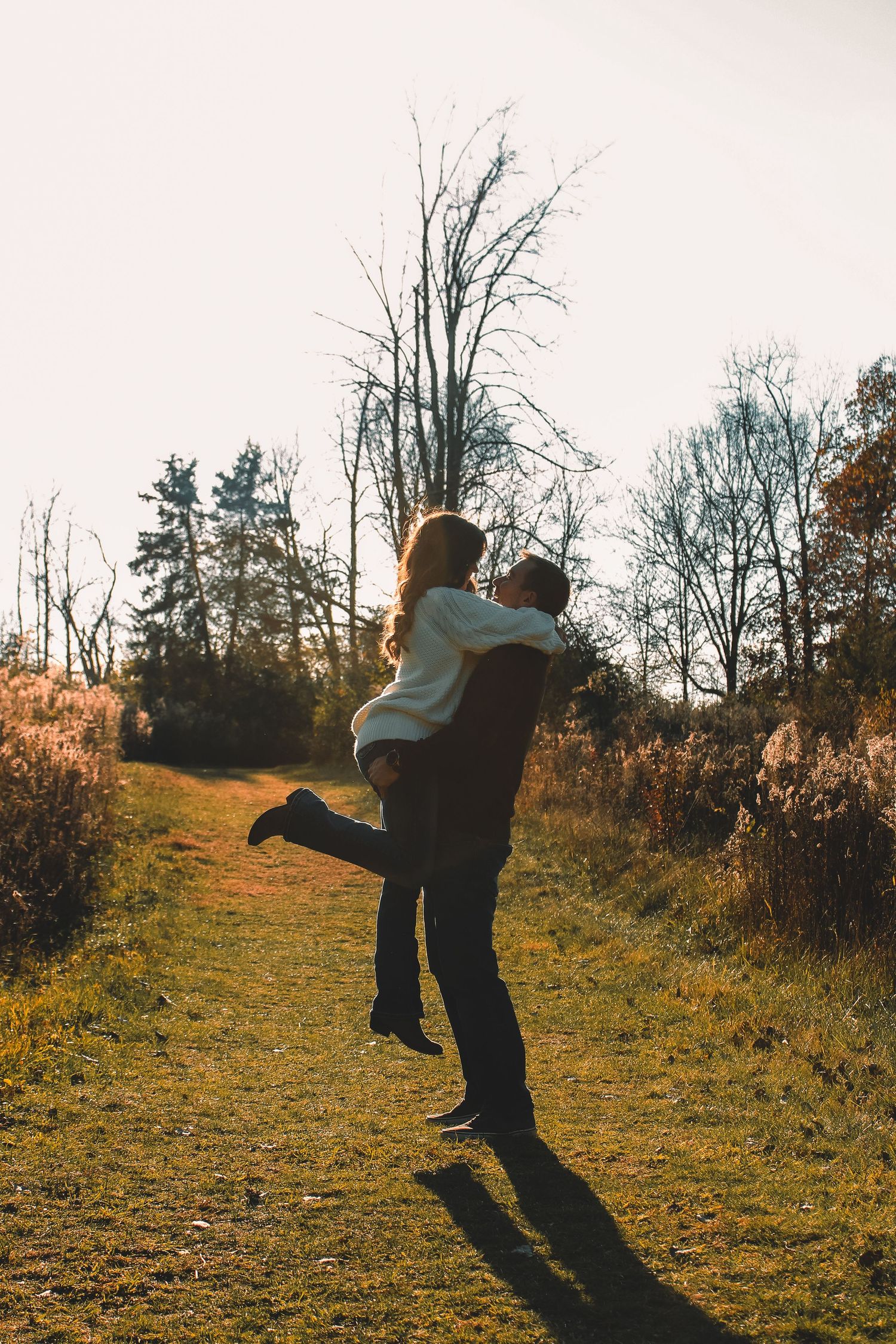 A couple embraces while silhouetted against bare trees and golden sunlight on a late autumn afternoon.