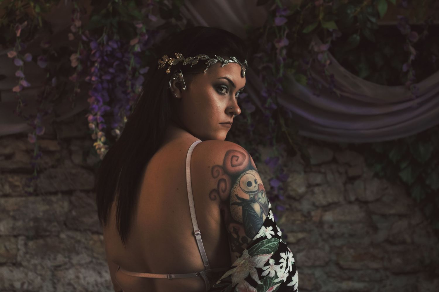 Artistic portrait featuring a model in shadow with an ornate skull tattoo against a dark stone wall with purple accents.