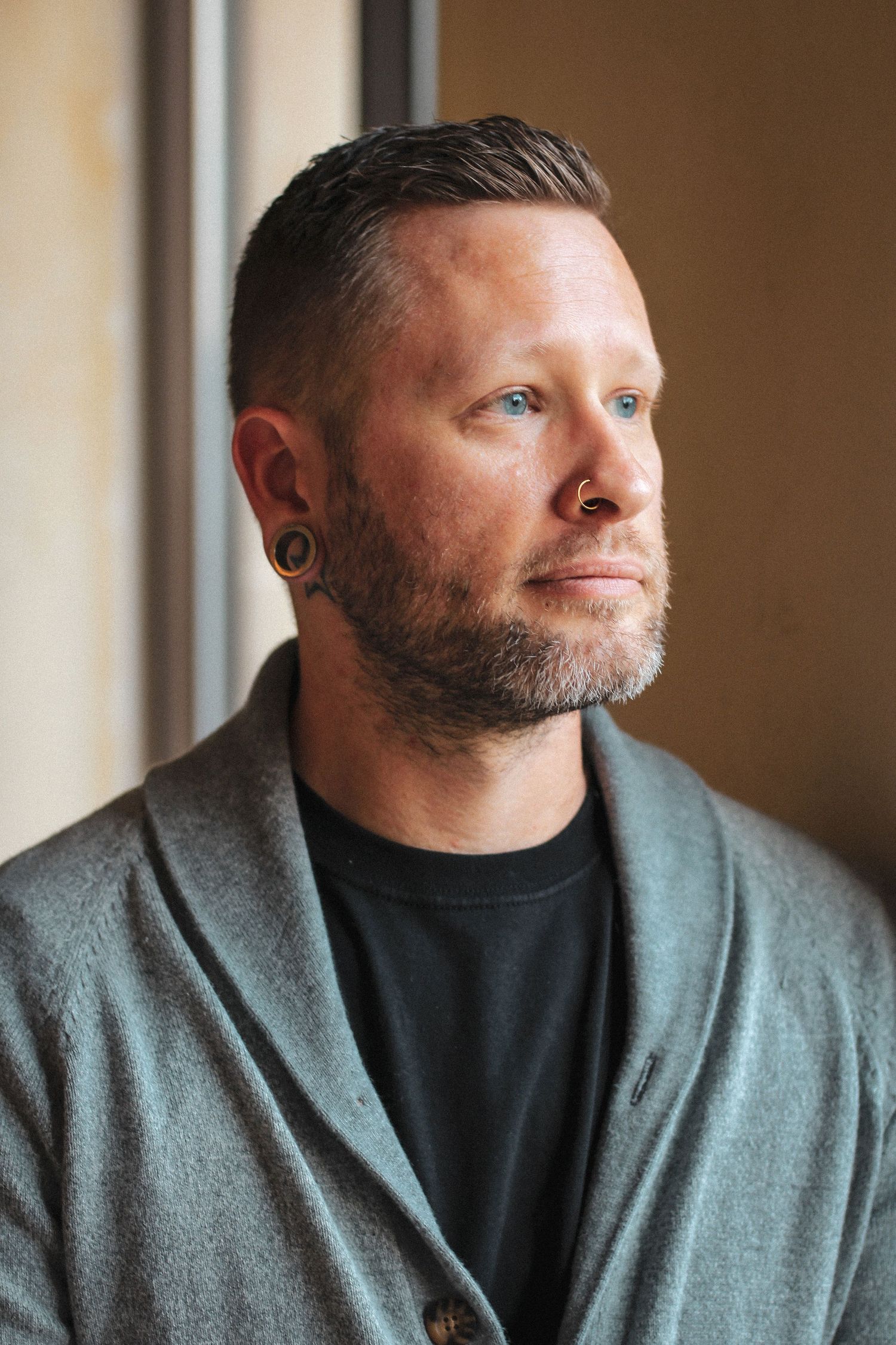 Portrait of person with beard wearing grey cardigan and black t-shirt looking thoughtfully through window in natural light.