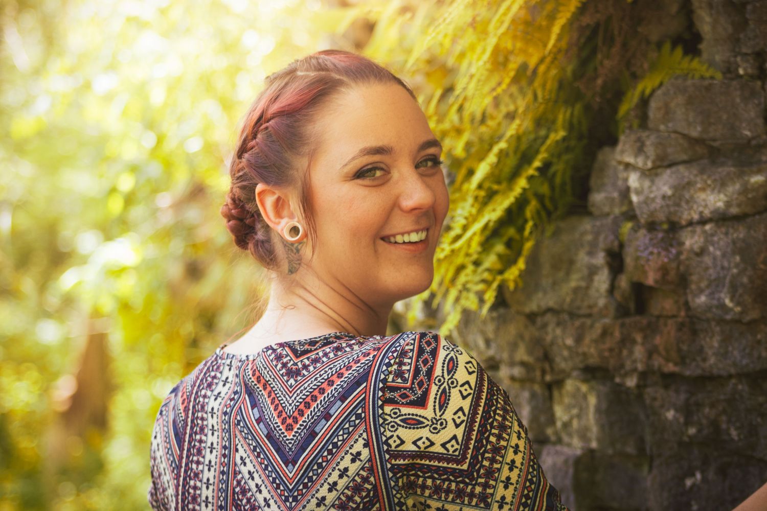A person wearing a patterned top smiles in soft golden sunlight near a stone wall with greenery above.