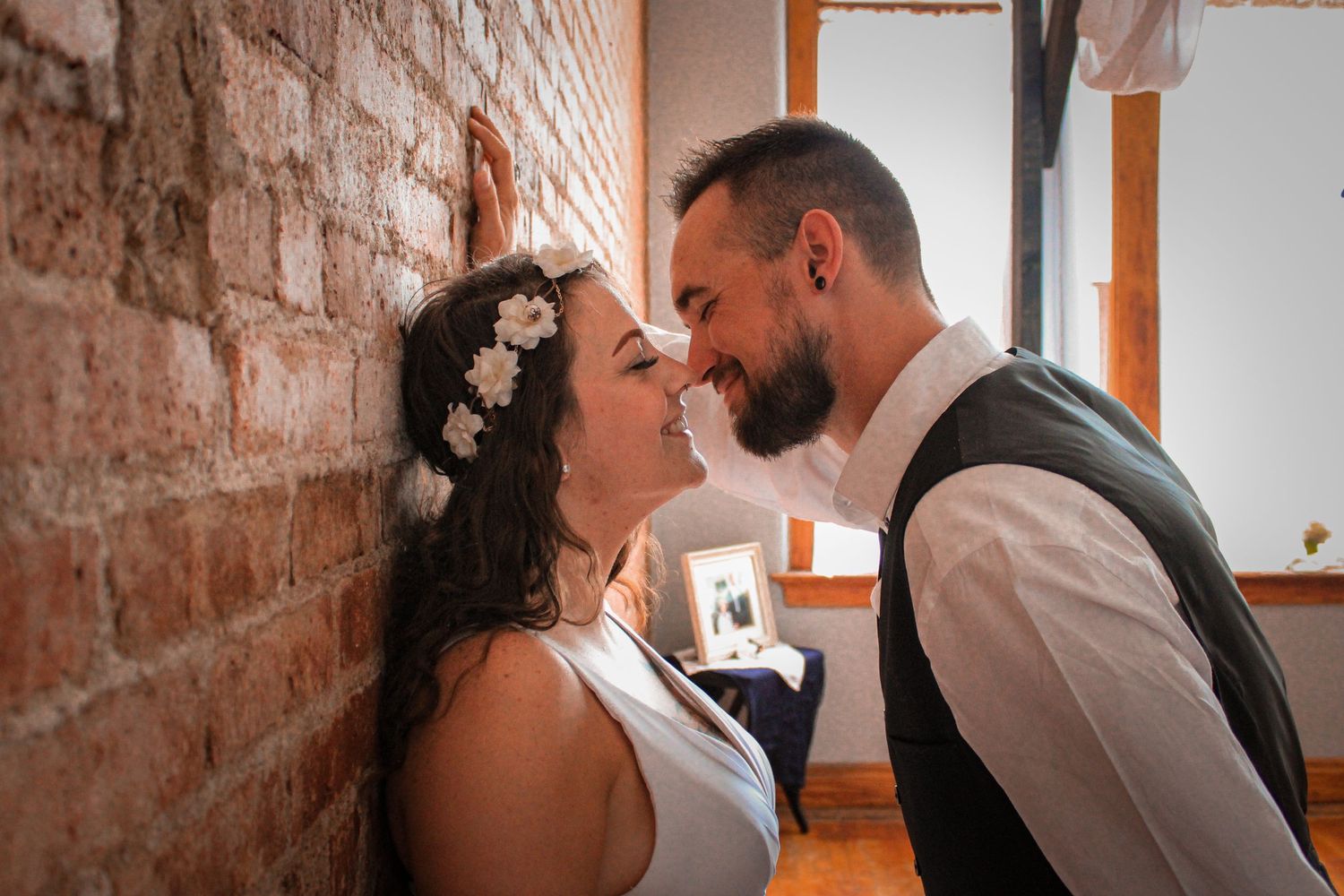 A happy wedding portrait of a couple sharing an intimate moment by a brick wall, bride wearing a floral crown.