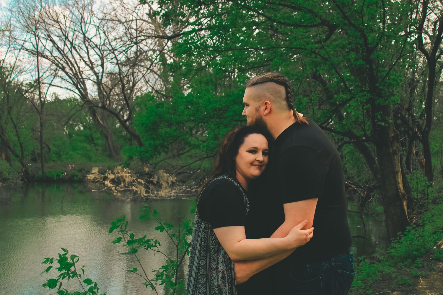 A couple embraces by a tranquil river surrounded by lush green trees on a peaceful spring afternoon.