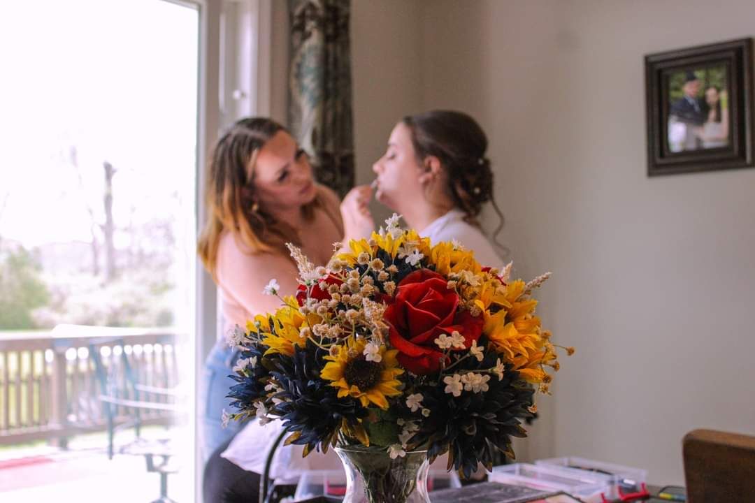 Bride getting makeup done by bridesmaid near window with colorful floral bouquet of sunflowers and roses in foreground.