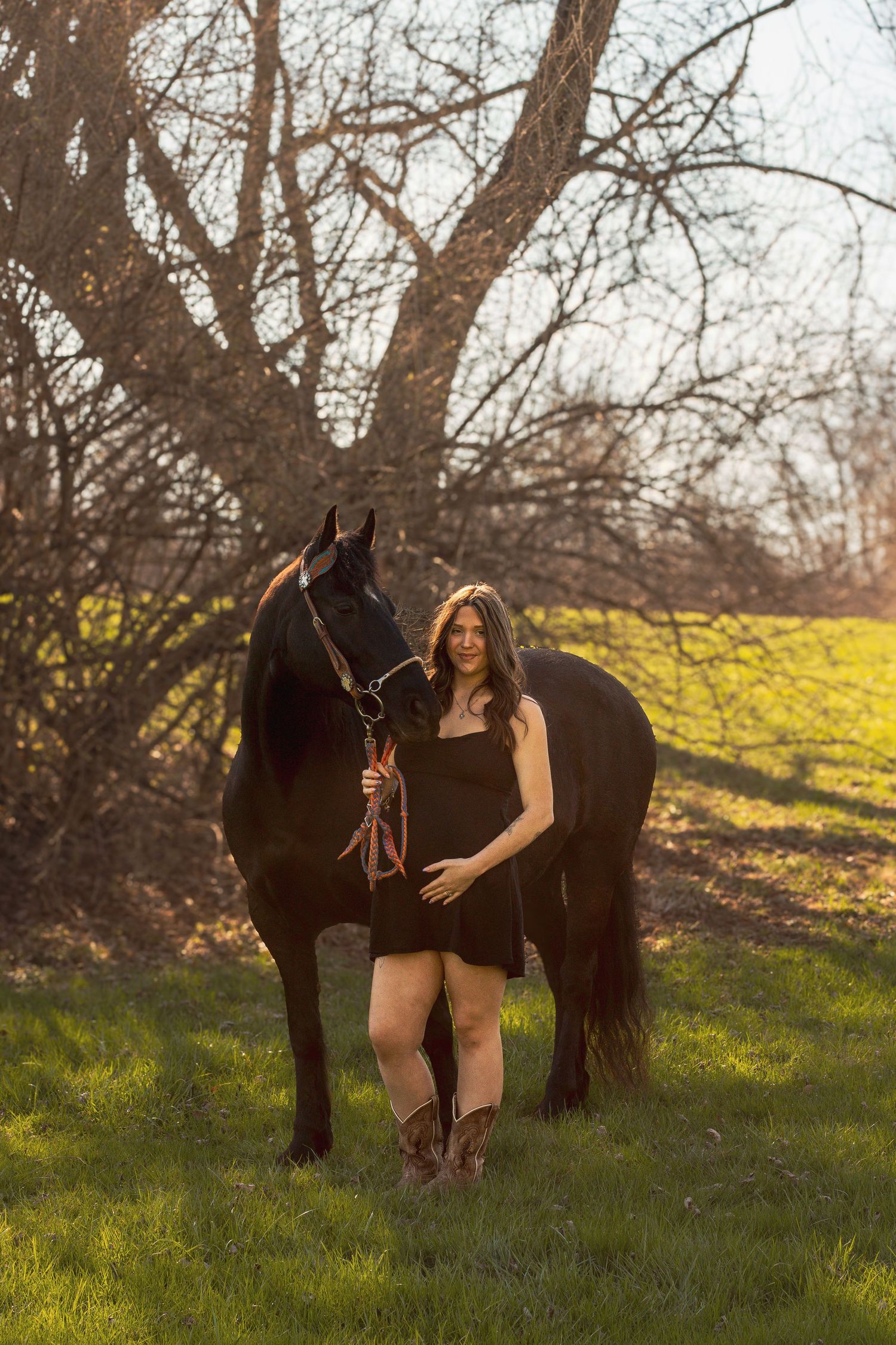 Woman in black dress and cowboy boots stands smiling beside a dark horse on green grass with bare trees behind.