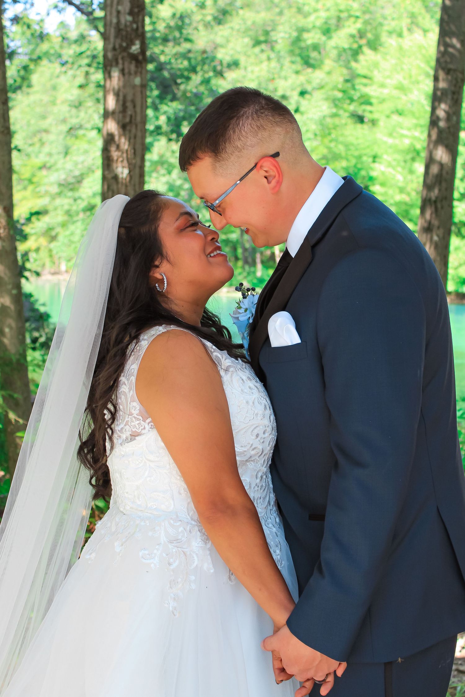 A bride in a white lace gown and groom in navy suit share an intimate moment in a wooded outdoor setting.