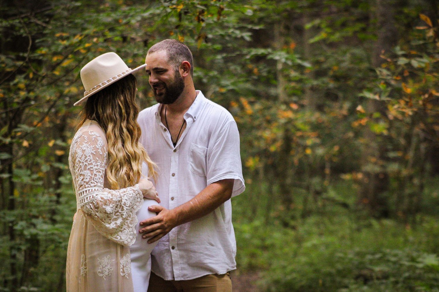 A romantic couple stands close together in a lush forest setting, wearing bohemian style clothing in neutral earth tones.