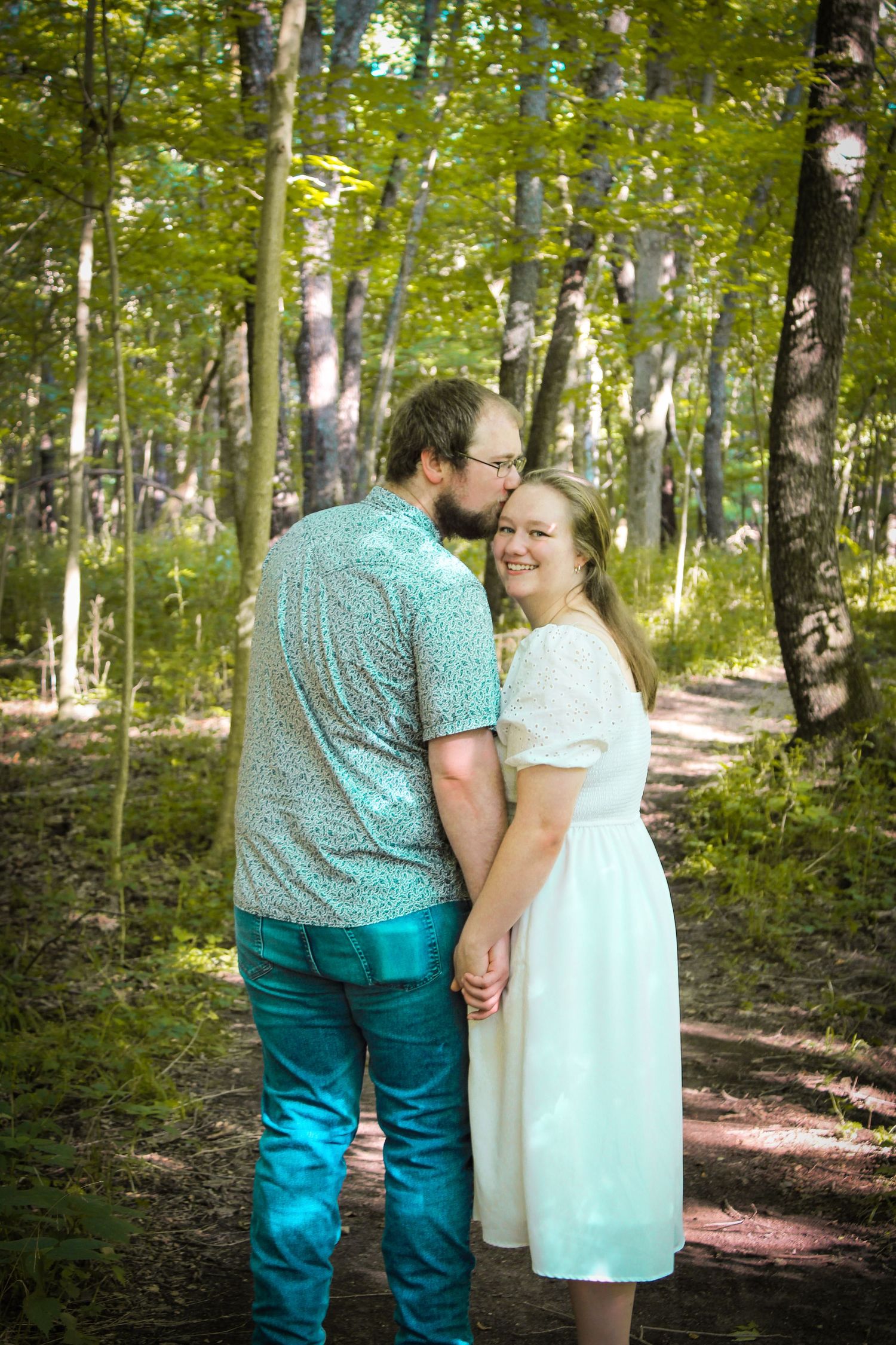 A couple shares a tender moment while standing on a forest path surrounded by green trees and dappled sunlight.