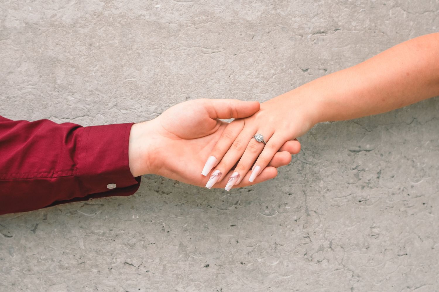 Two hands reaching out to hold each other against a gray concrete background, one wearing a burgundy sleeve.