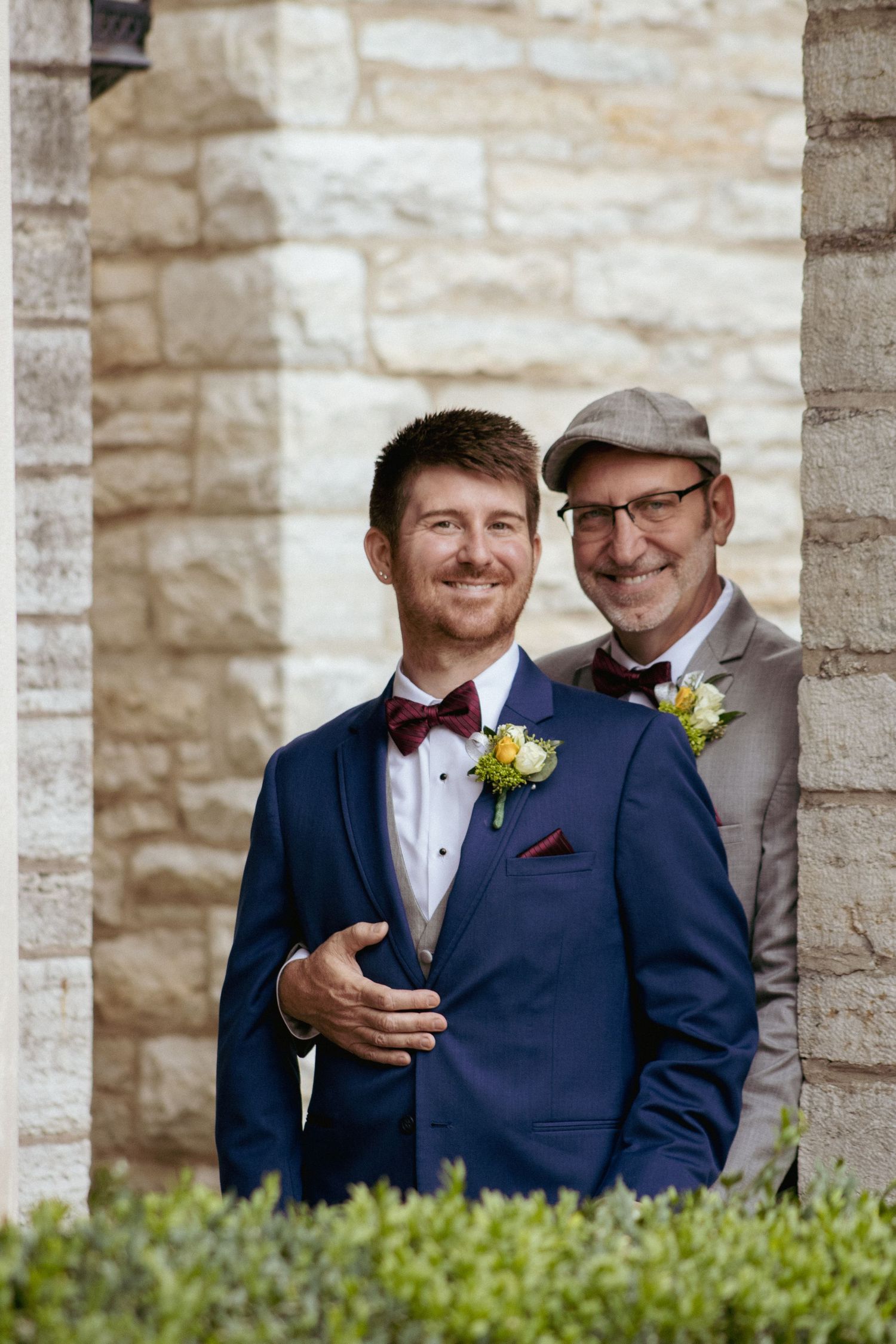 Two people wearing suits and boutonnieres pose together at a formal event with stone wall and greenery backdrop.