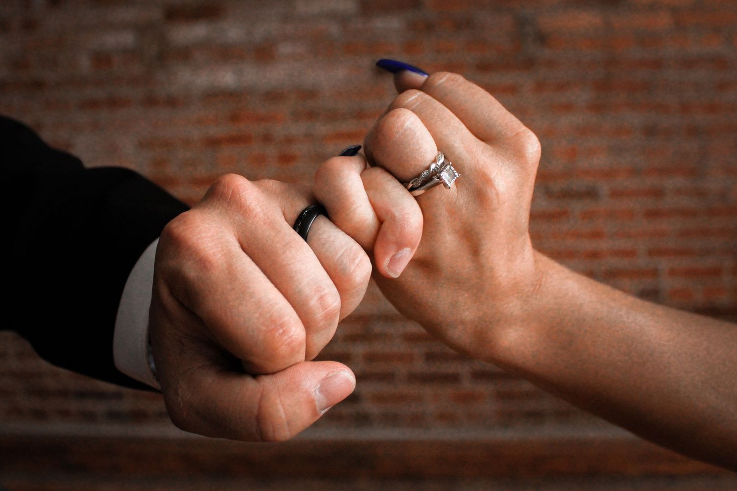 Two hands making a pinky promise gesture against a brick wall background, with one hand wearing a silver ring.