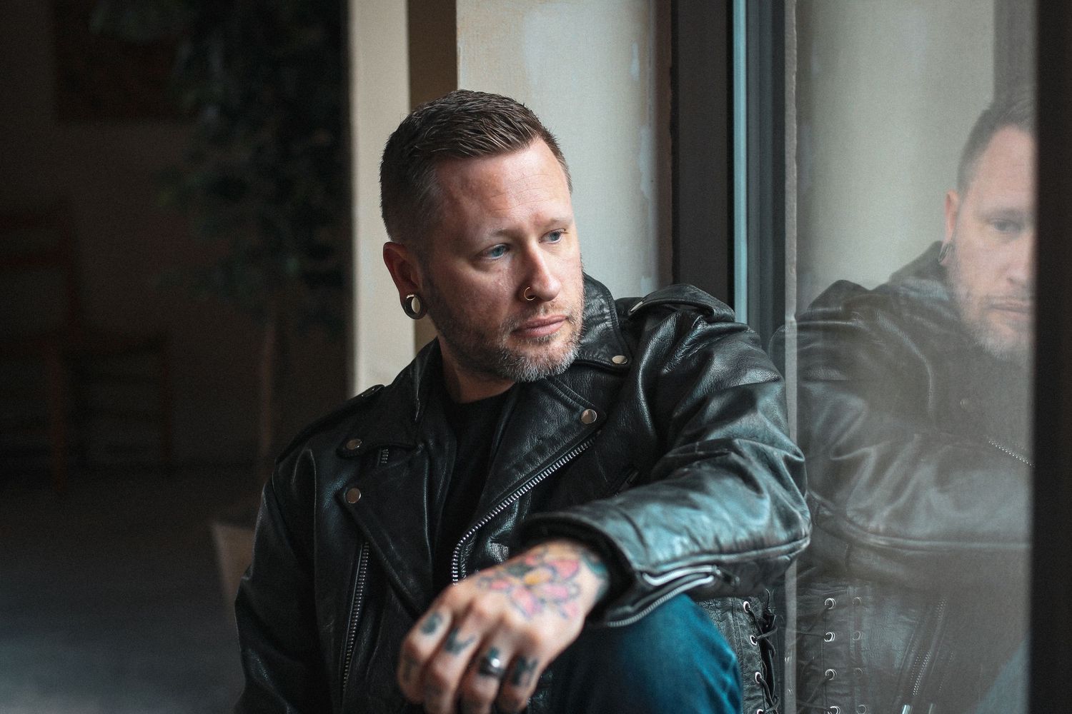 Person in black leather jacket sits contemplatively by window with reflection visible in moody lighting.