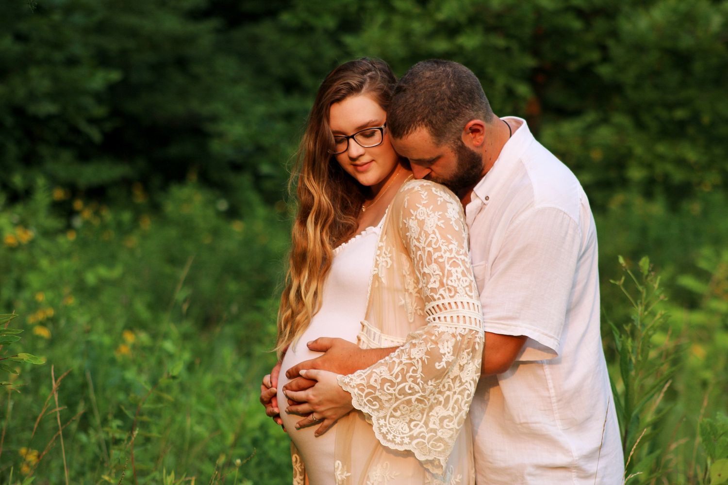 A romantic couple embraces in a lush green meadow wearing white clothing during a sunny outdoor photo session.