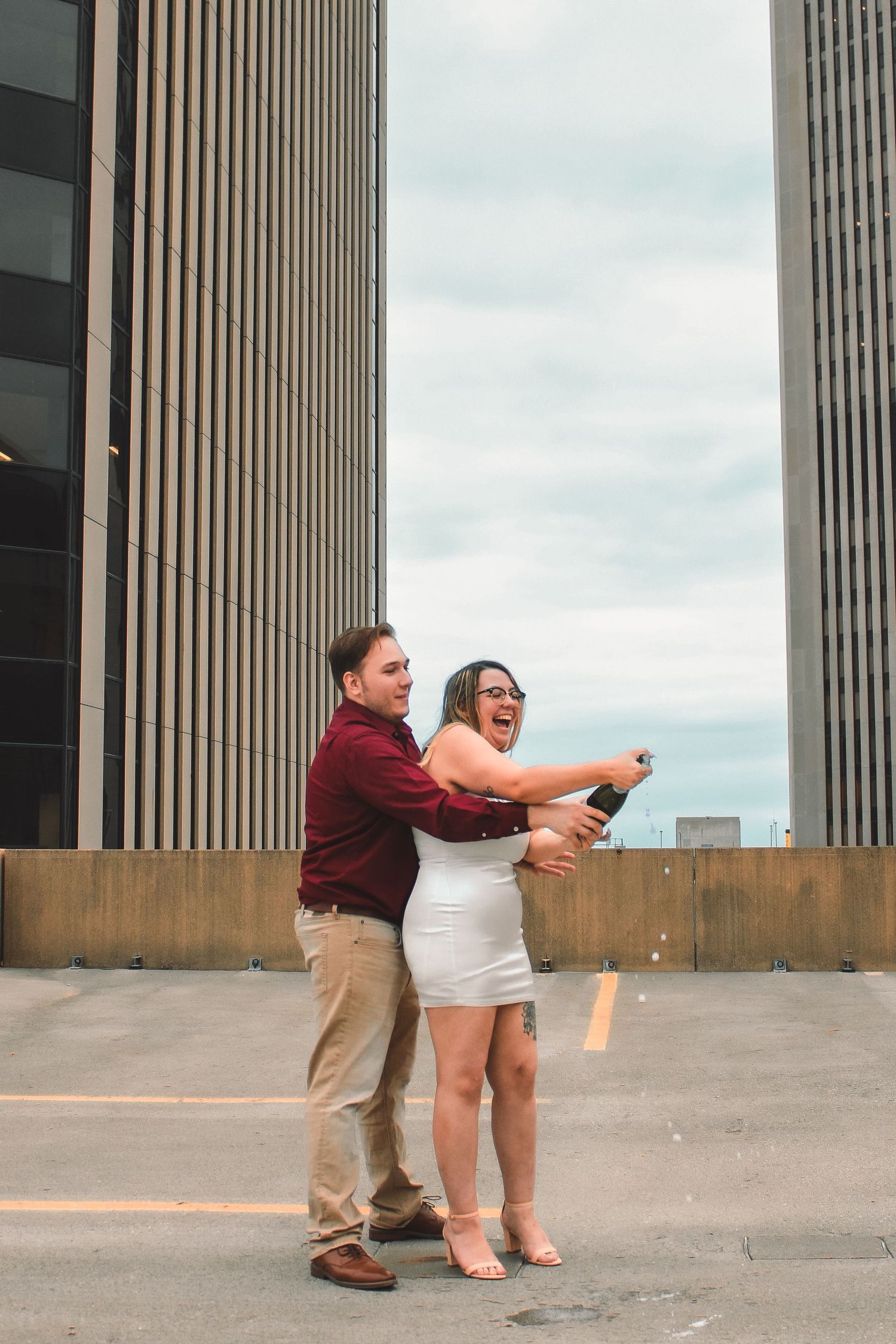 A couple embraces and laughs together on a rooftop parking deck with tall office buildings in the background.