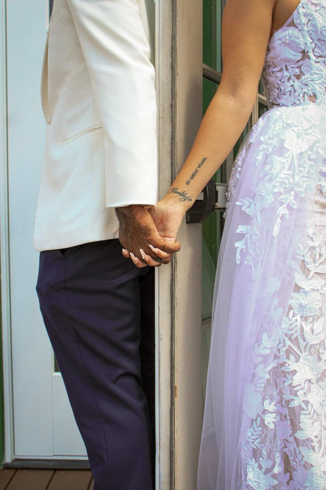 Couple holding hands, groom in white blazer and navy pants, bride in lavender lace gown with wrist tattoo visible.