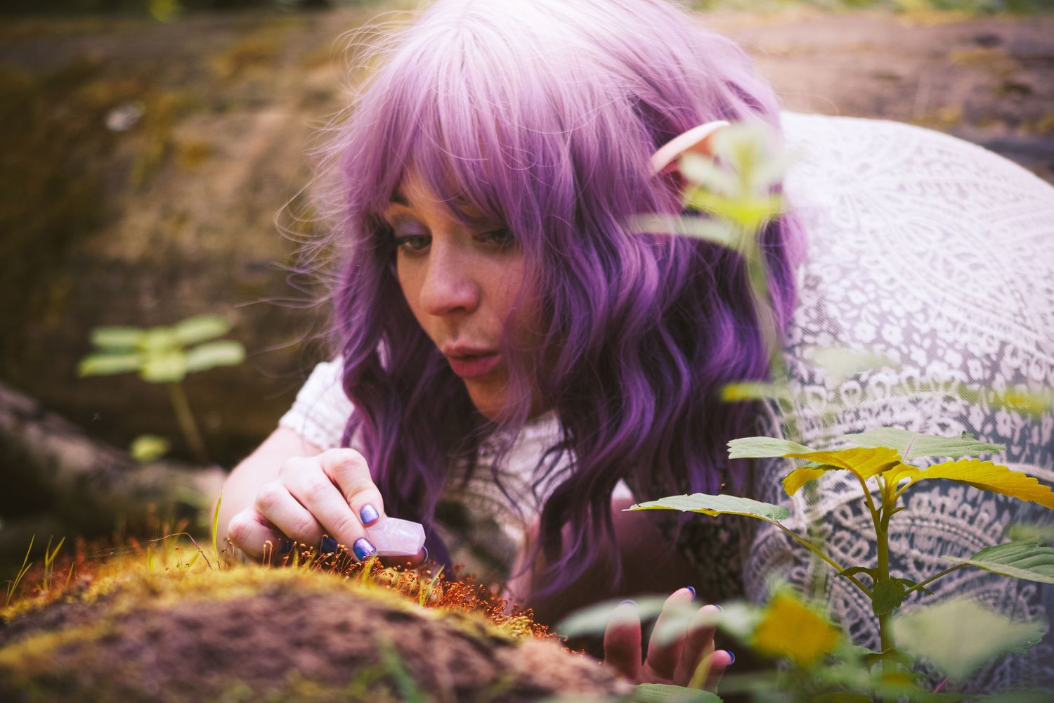 A figure with vibrant purple hair lying on the ground in nature looking closely at a crystal in the moss.