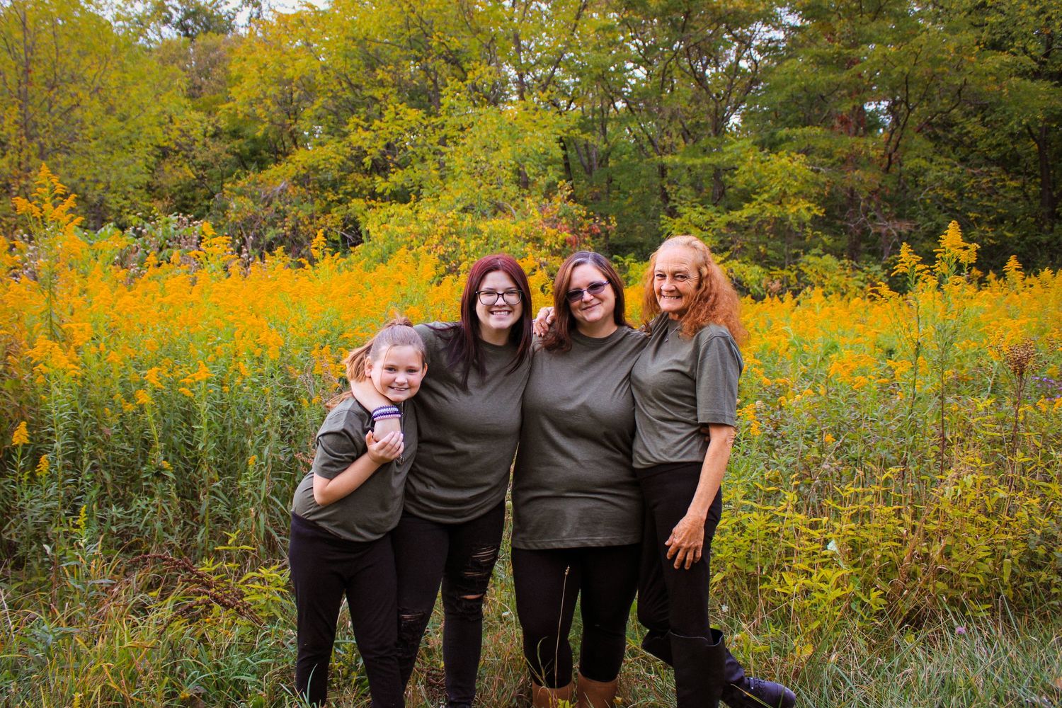A group stands close together smiling in a field of vibrant yellow wildflowers with autumn trees in the background.