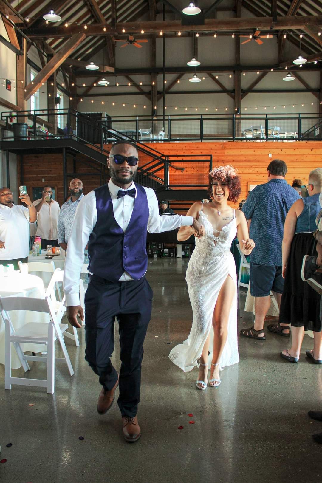 Bride and groom make grand entrance at wedding reception in rustic barn venue with exposed wood beams and string lights.