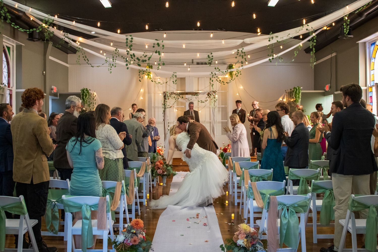 Newlyweds share a kiss at the end of the aisle during an indoor wedding ceremony with floral arch and string lights.