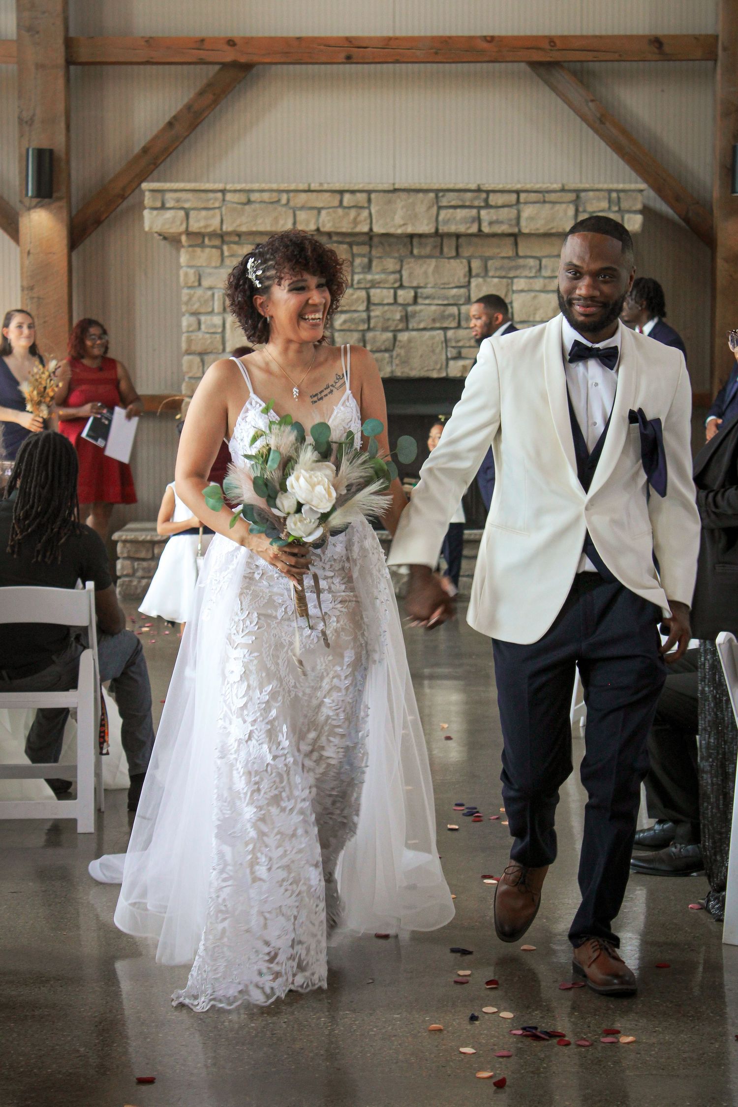 A bride in white lace gown and groom in white tuxedo jacket joyfully walk together at their wedding ceremony.