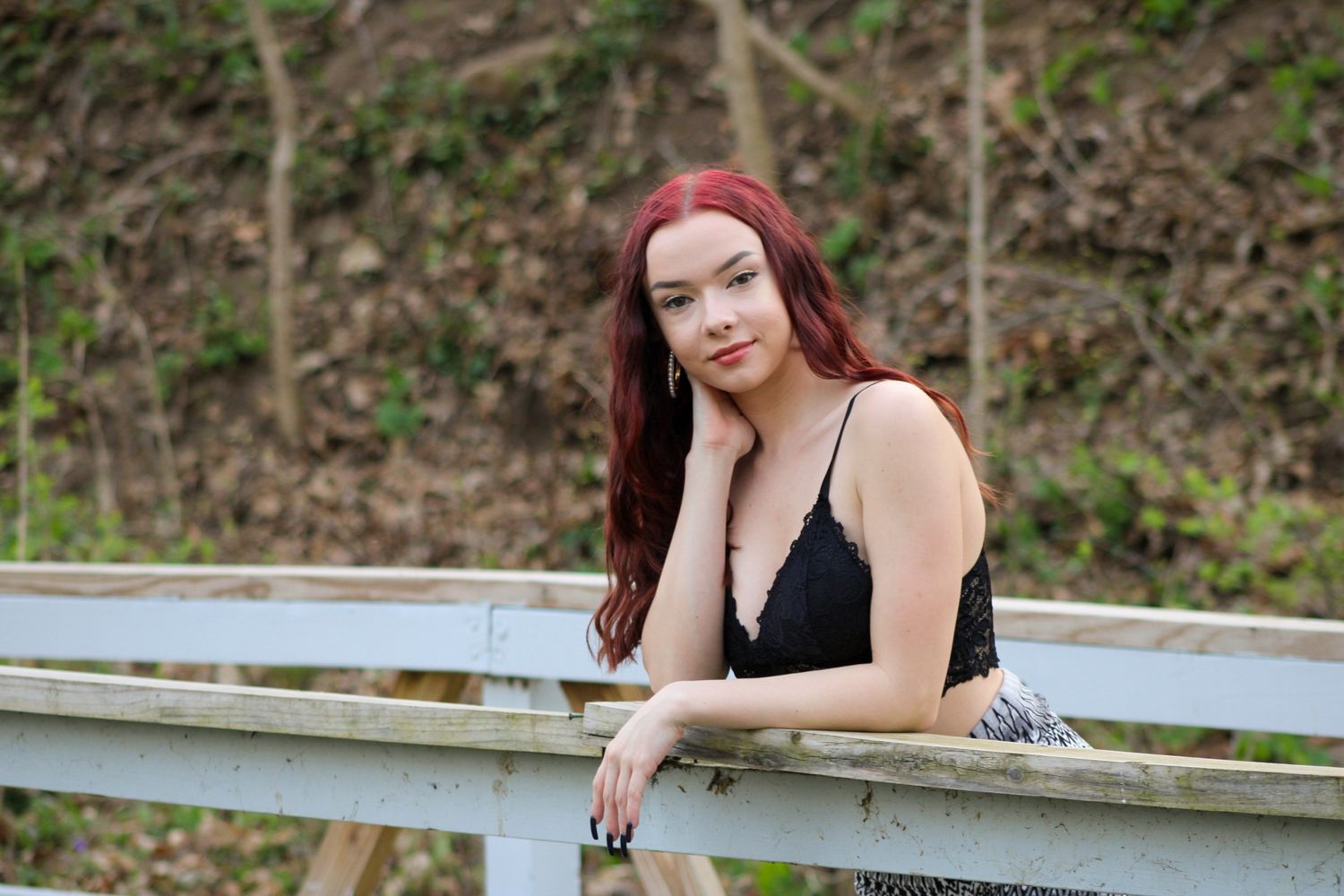 Person with long red hair in black top leaning on wooden railing outdoors on sunny day.