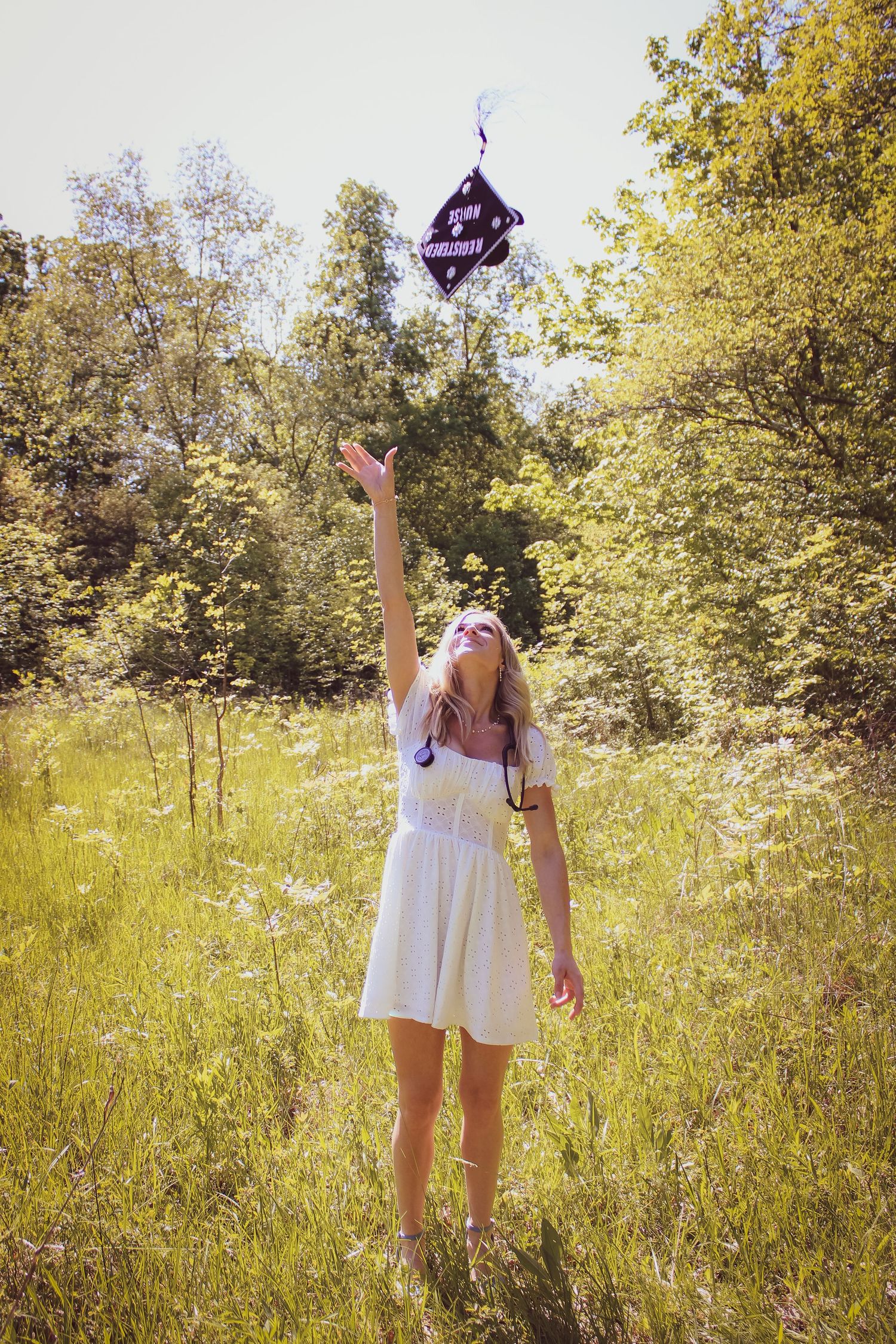 A figure in a white dress reaches up towards a floating purple lantern in a sunny meadow surrounded by trees.