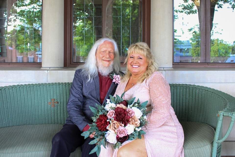 Smiling couple in wedding attire sitting on bench, bride holding bouquet of red and pink flowers.