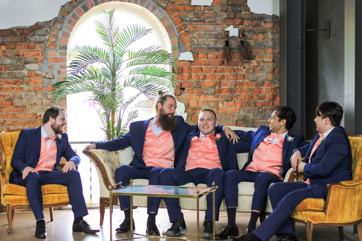 A group wearing matching suits sitting together on chairs against an exposed brick wall with an arched window.