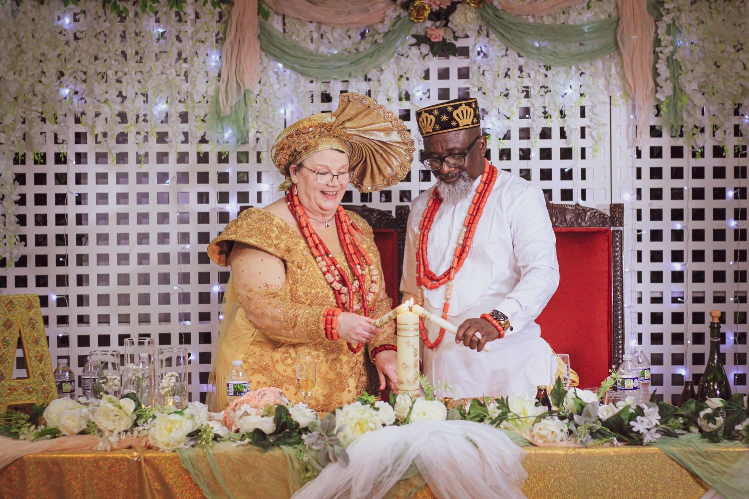 A festive wedding ceremony with couple in traditional gold and white attire seated at a decorated table with floral arrangements.
