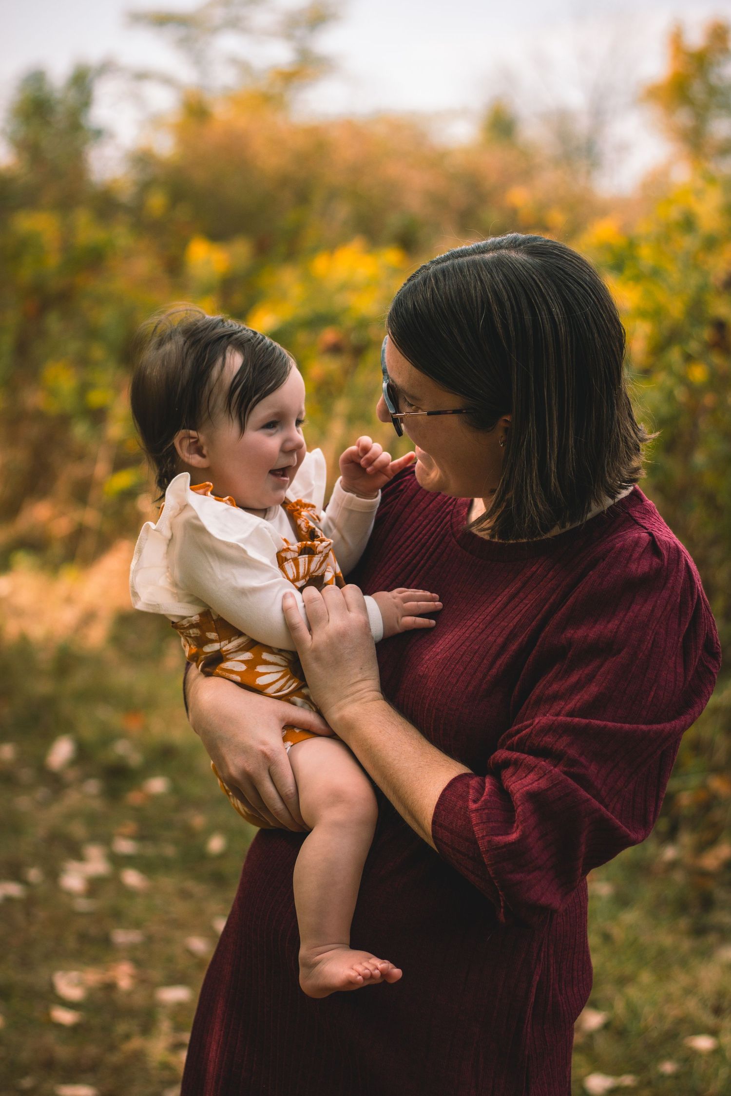 A tender moment captured outdoors in autumn as an adult holds a laughing baby wearing a white top against fall foliage.